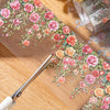 A pair of scissors is cutting a floral vine transparent PET washi tape featuring pink and orange roses onto a wooden surface.