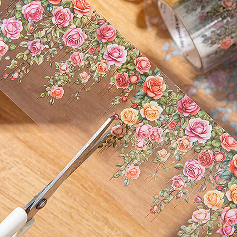 A pair of scissors is cutting a floral vine transparent PET washi tape featuring pink and orange roses onto a wooden surface.