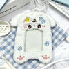 A white plush photocard holder with a soft cover featuring cute star and cube decorations, resting on a checkered cloth.