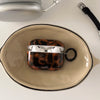 A tortoiseshell patterned protective case for AirPods rests in a light ceramic bowl.