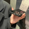 A hand holds a black phone case featuring gothic floral art on an iPhone against a dark background.