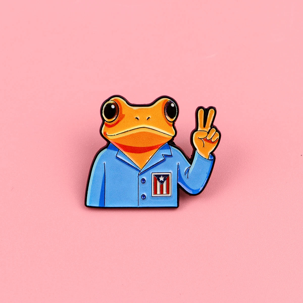 An enamel pin featuring a frog wearing a blue shirt with a patriotic design making a peace sign.
