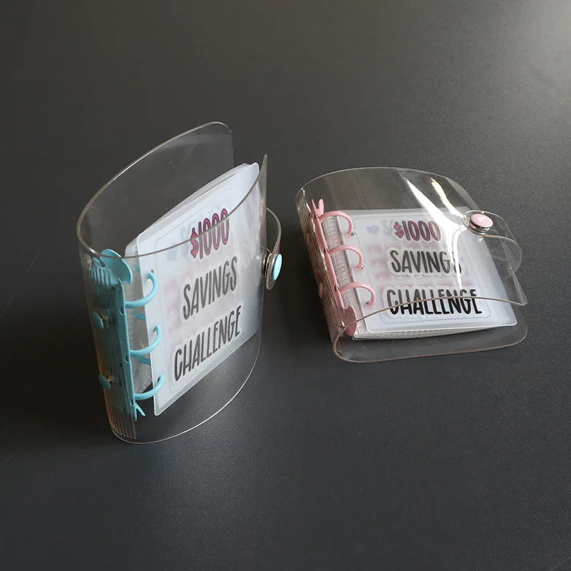 A clear plastic binder for a savings challenge with pink and teal accents on a dark surface.