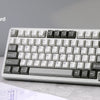 A white B75 PRO gasket mechanical keyboard is shown on a desk next to some blueprints.