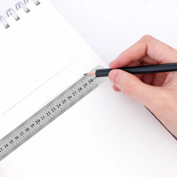 Une main tient un crayon noir contre une règle graduée en acier inoxydable sur une feuille de papier blanc.