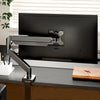 A silver adjustable monitor arm is mounted to a black computer monitor on a dark desk.