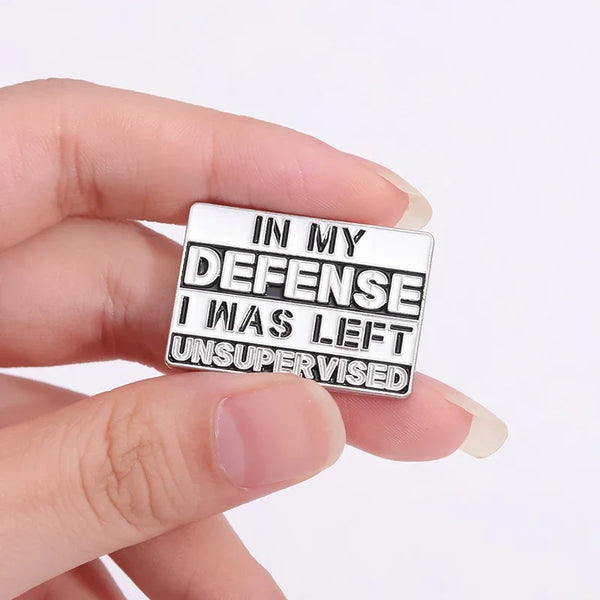Silver enamel pin with text 'In My Defense I Was Left Unsupervised' held between fingers.