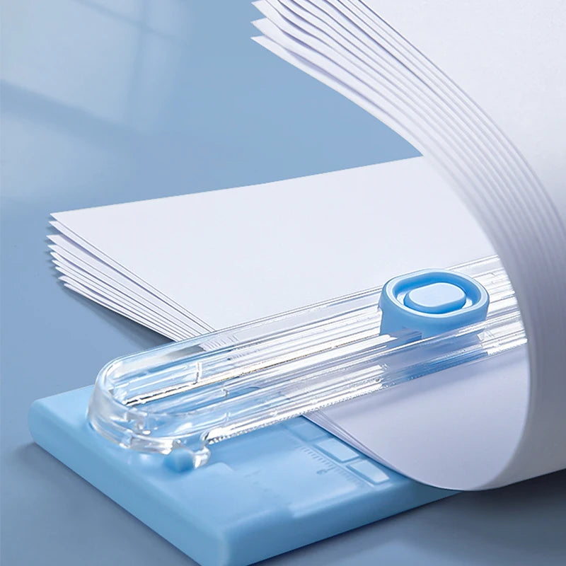 A portable manual paper cutter trimmer with a clear plastic blade is shown resting on a light blue base.