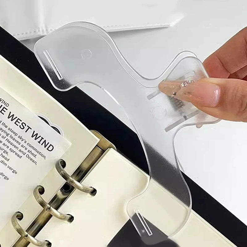 A clear plastic book page clip is shown being held by a hand, positioned over a white notebook.