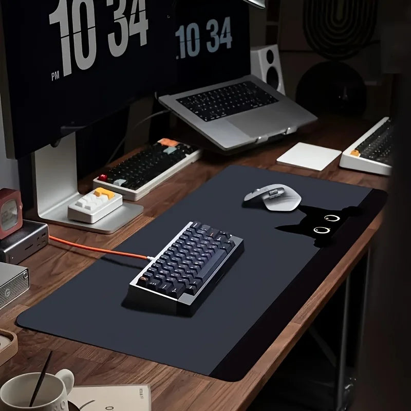 A large black non-slip gaming desk mat featuring a cute cat mouse design placed on a wooden desk with computer equipment.