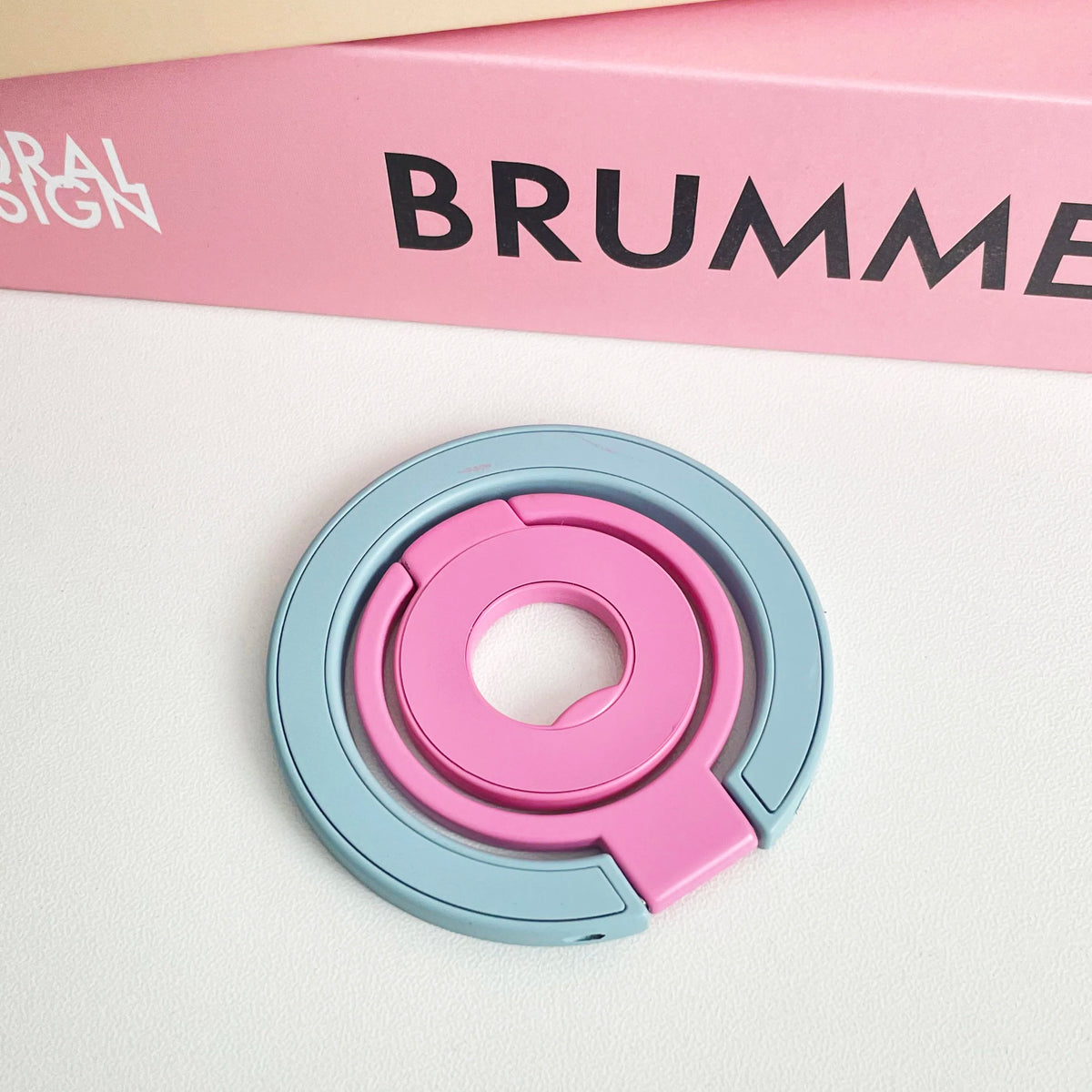A pink and blue magnetic phone grip and stand with a foldable ring resting on a white surface.