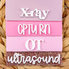 A set of pink acrylic badge holders with white text spelling out XRAY, CTURN, OT, and ultrasound.