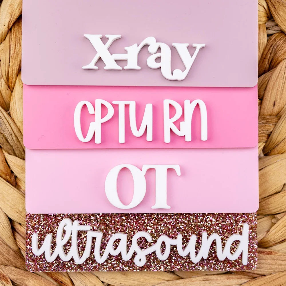 A set of pink acrylic badge holders with white text spelling out XRAY, CTURN, OT, and ultrasound.