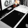 A black extra large gaming mouse pad is displayed on a white desk next to a monitor and a desk lamp.