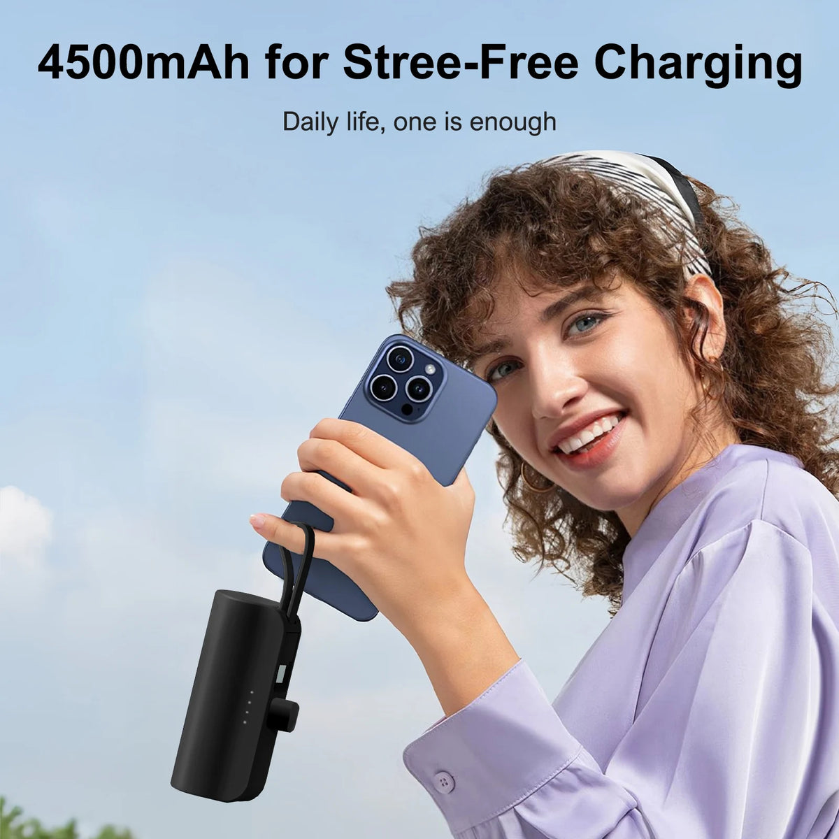 A young woman is using a compact black power bank to charge a smartphone outdoors.
