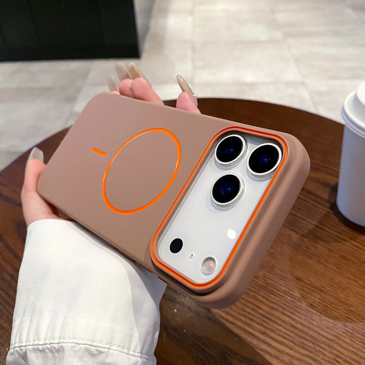 A hand holds a beige magnetic wireless charging phone case with a clear back and three camera lenses.