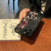 A dark floral art phone case for an iPhone resting on a wooden table next to a notebook.