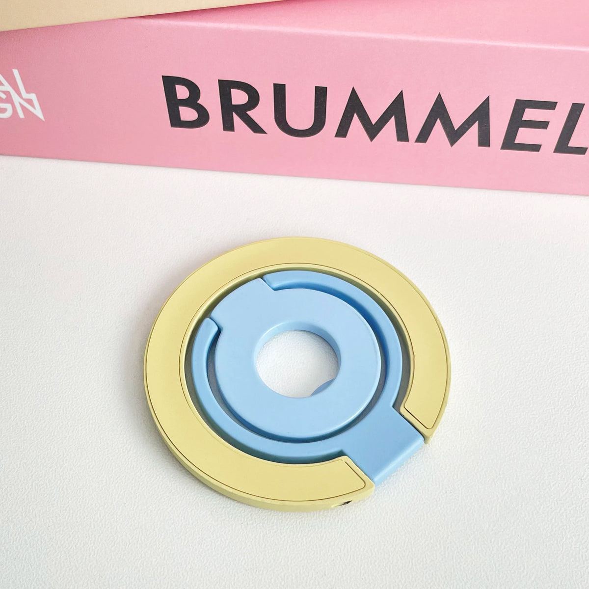 A light blue and yellow magnetic phone grip and stand with a foldable ring resting on a white surface.