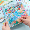 A hand holds an open sticker book featuring a cute snack shop scene with various food items and colorful decorations.