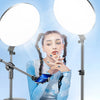 A woman holds up a blue smartphone in front of two bright LED ring lights mounted on tripods.