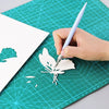 A hand writes on a teal cutting mat, drawing a leaf design with a white pen and grid lines.