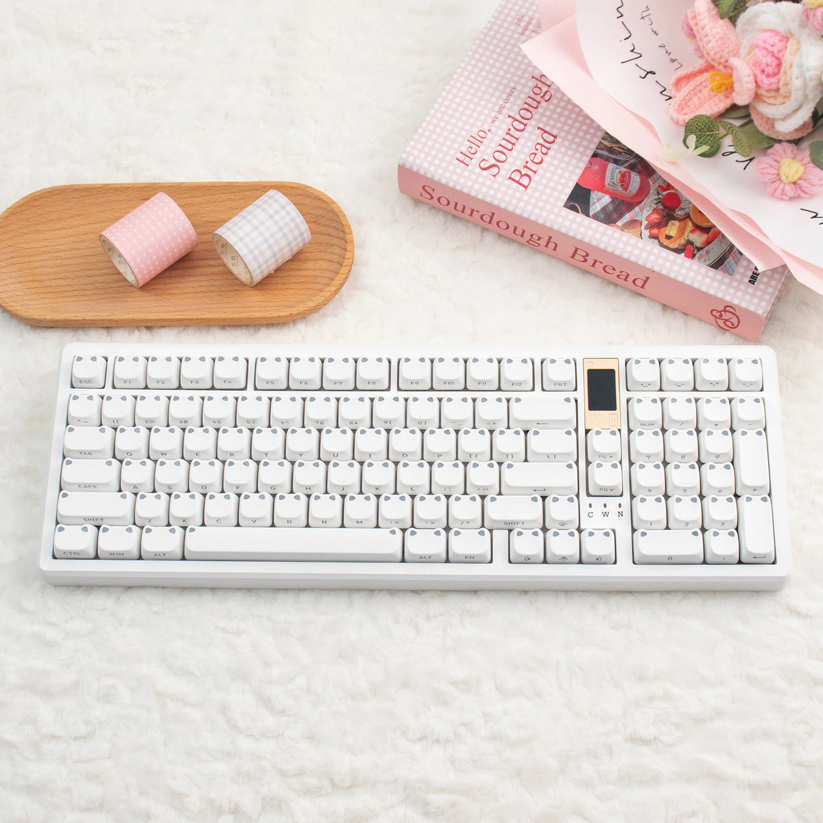 Eine weiße mechanische Tastatur neben rosa Brot und dekorativen Blumen auf weißem Untergrund.