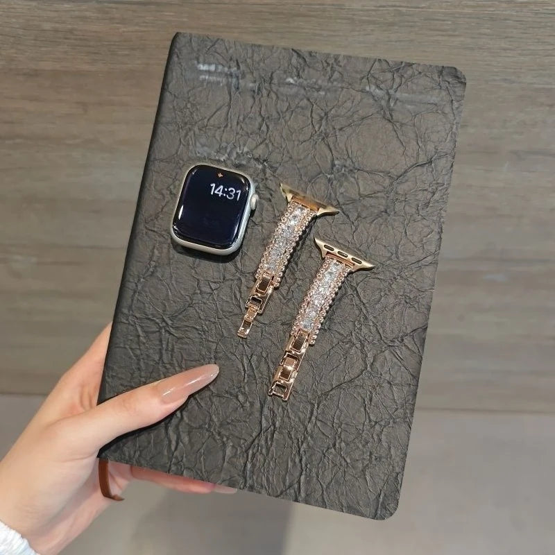 A close-up of a black smartphone and two crystal-embellished gold watch straps resting on a dark gray leather surface.