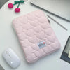 A pink quilted laptop sleeve is shown on a white table next to a silver laptop and a white wireless mouse.