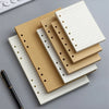 Una pila de resmas sueltas de cuaderno beige y blanco con encuadernación dorada y un bolígrafo negro sobre una superficie gri