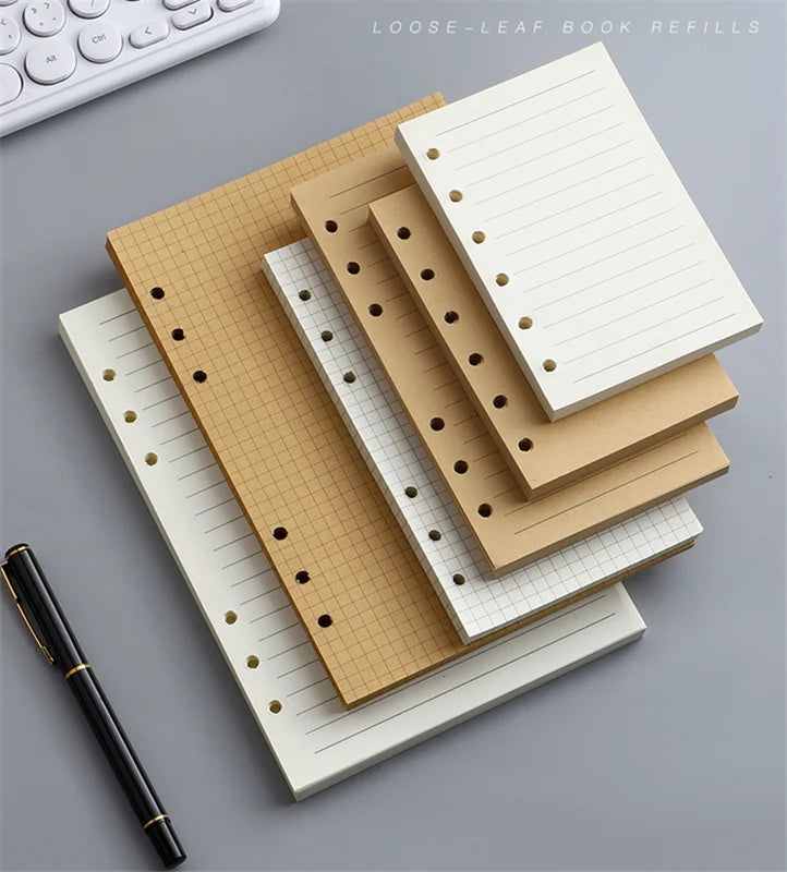 Una pila de resmas de blocs de papel suelto beige y blanco con encuadernación dorada y un bolígrafo negro sobre una superfici