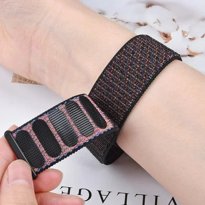 A close-up of a black nylon loop watch strap with pink and blue accents being held by a hand.