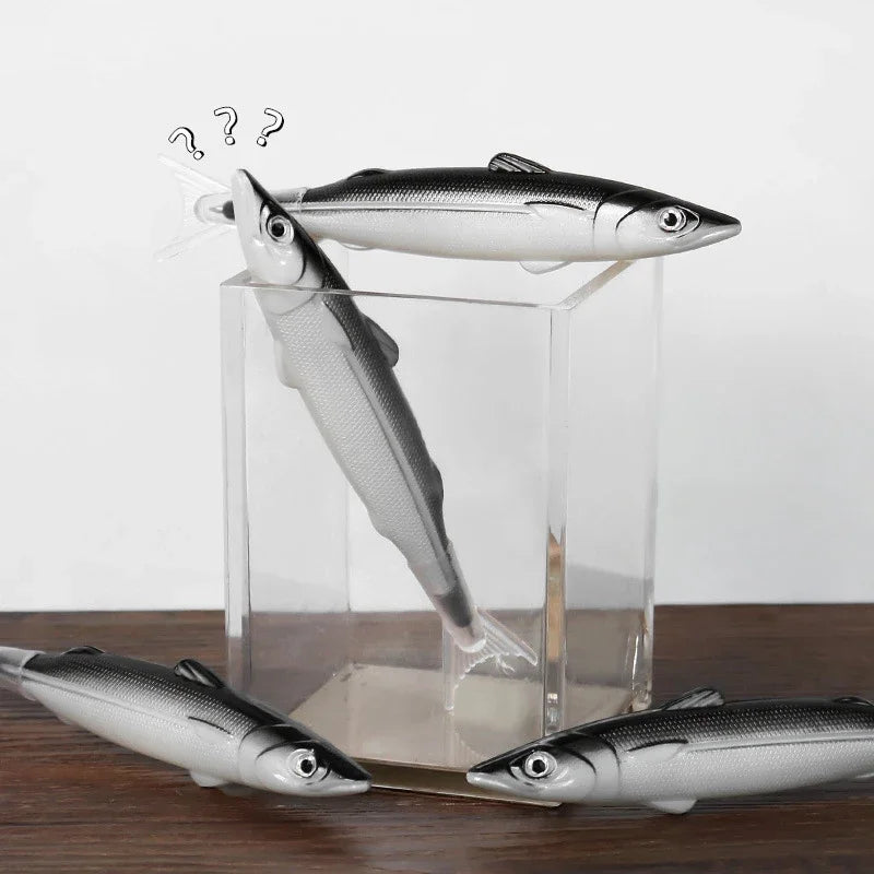A clear glass display box holds three silver fish-shaped ballpoint pens on a wooden surface.