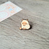 Un pequeño pin de esmalte beige claro en forma de un gato tierno sentado sobre una superficie de madera.