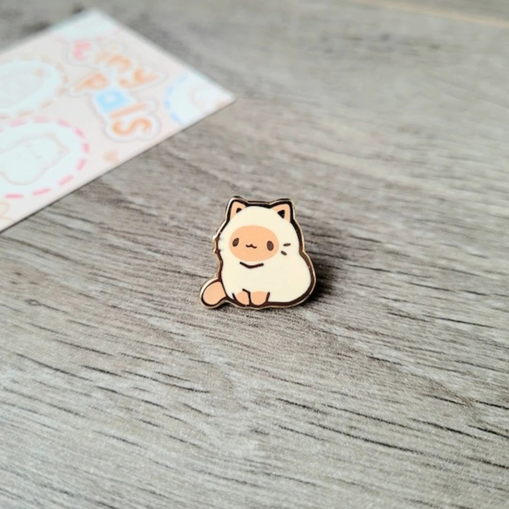 Un pequeño pin de esmalte beige claro en forma de un gato tierno sentado sobre una superficie de madera.