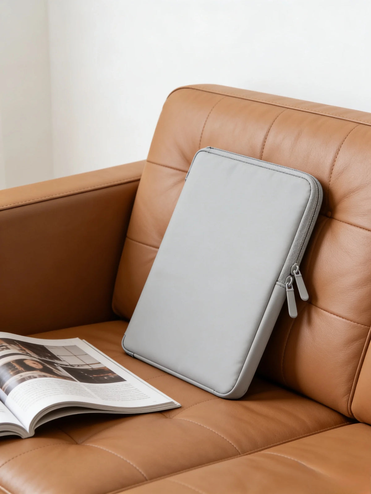 A gray waterproof PU leather laptop sleeve resting on a brown leather sofa next to an open magazine.