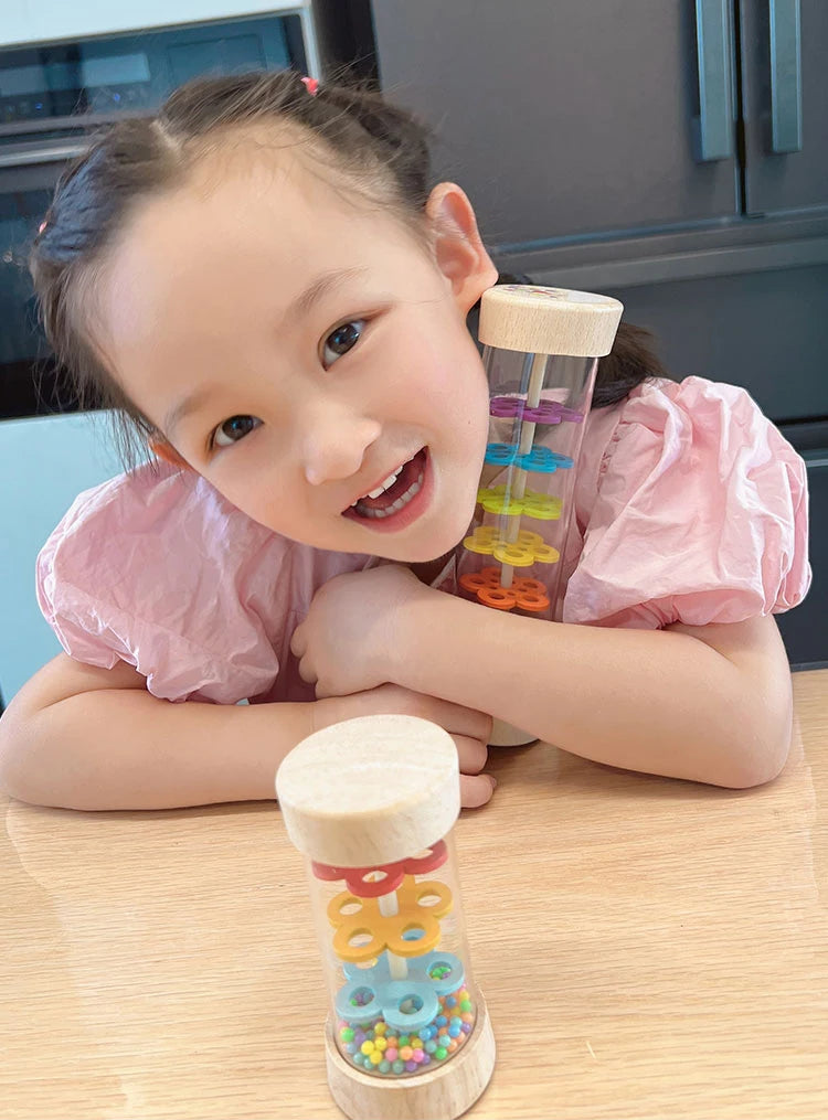 A young girl holds a wooden rain sound tube musical instrument filled with colorful beads and a wooden stopper.