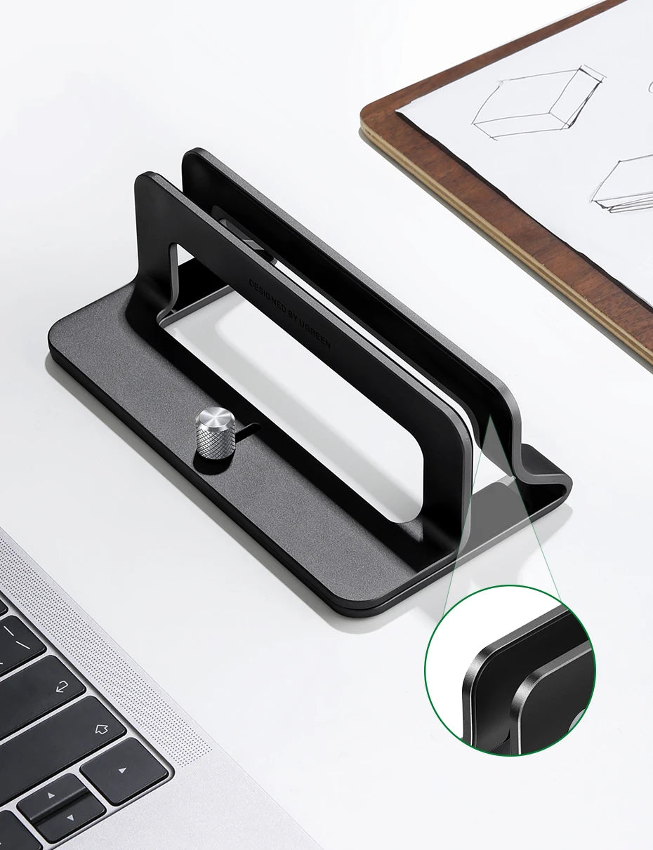 A black vertical laptop stand designed to hold a MacBook or tablet, shown next to a laptop keyboard.