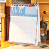 Calendario de escritorio vertical para mayo con cuadrícula mensual y acentos florales decorativos, colocado sobre una superfi