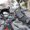 A hand wearing a black glove holds a smartphone mounted on a motorcycle handlebar, showing a universal phone mount.