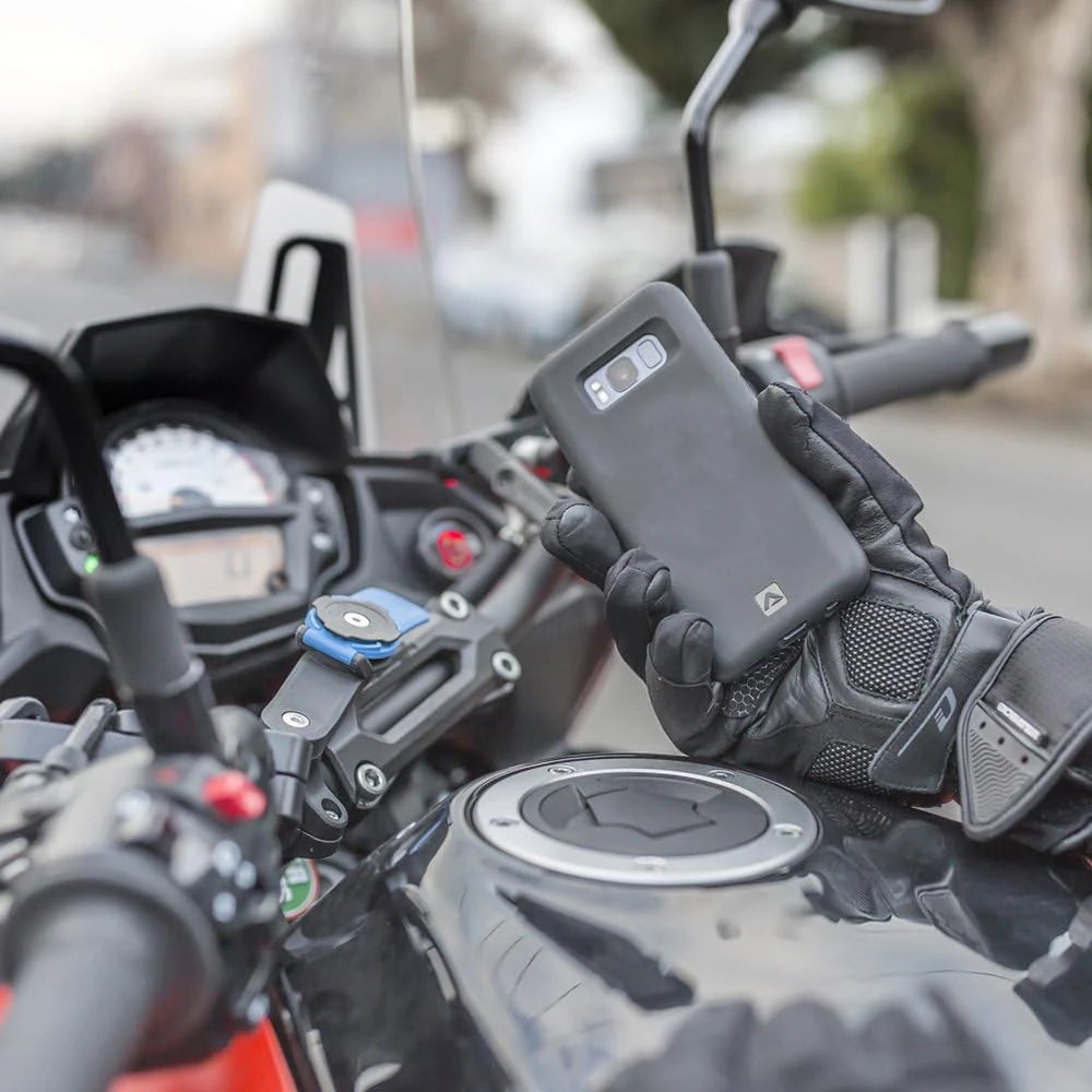 A hand wearing a black glove holds a smartphone mounted on a motorcycle handlebar, showing a universal phone mount.