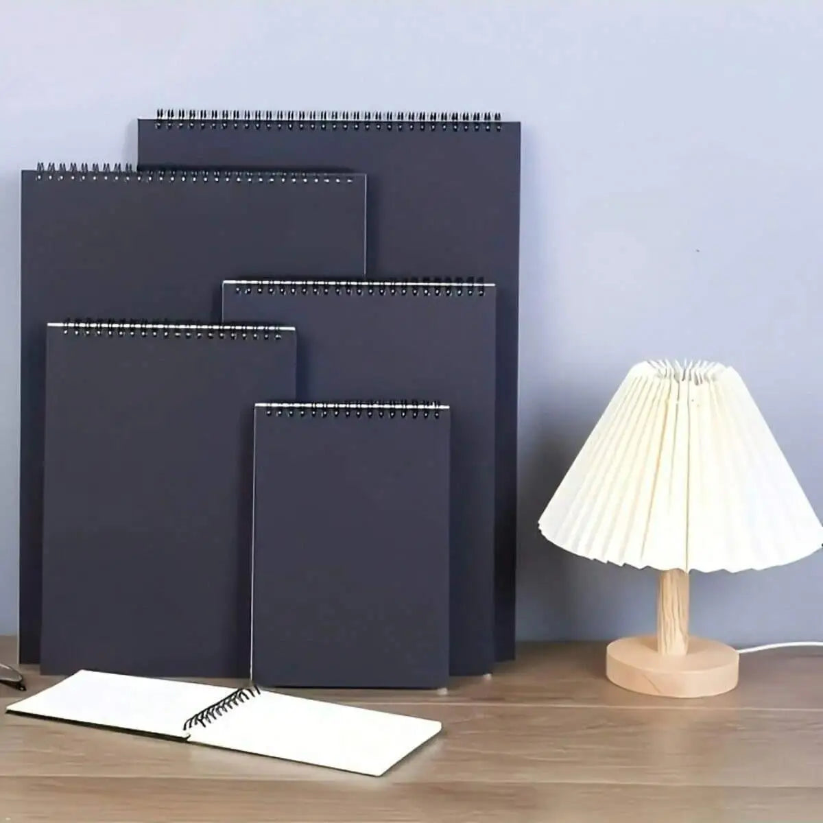 A set of dark gray spiral-bound sketchbooks and a wooden table lamp are displayed on a light surface.