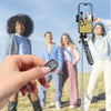 A hand holds a black multifunctional selfie stick tripod with a Bluetooth remote, set against a background of people outdoors
