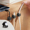 A hand is shown using magnetic cable organizer clips to secure a white charging cable on a wooden surface.
