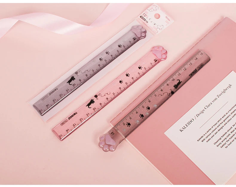 A pink plastic measuring tool with a cat paw design is shown next to a pink notebook.