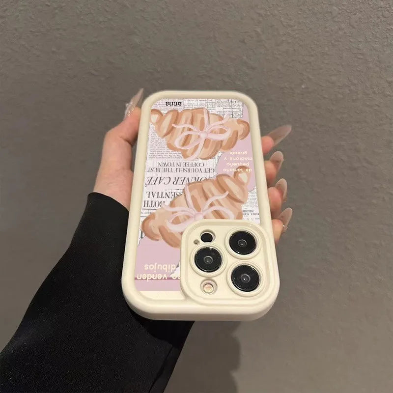 A beige phone case with a croissant newspaper pattern is held in a hand against a gray wall.