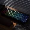 A black mechanical keyboard with colorful RGB lighting is displayed on a wooden surface next to a window blind.