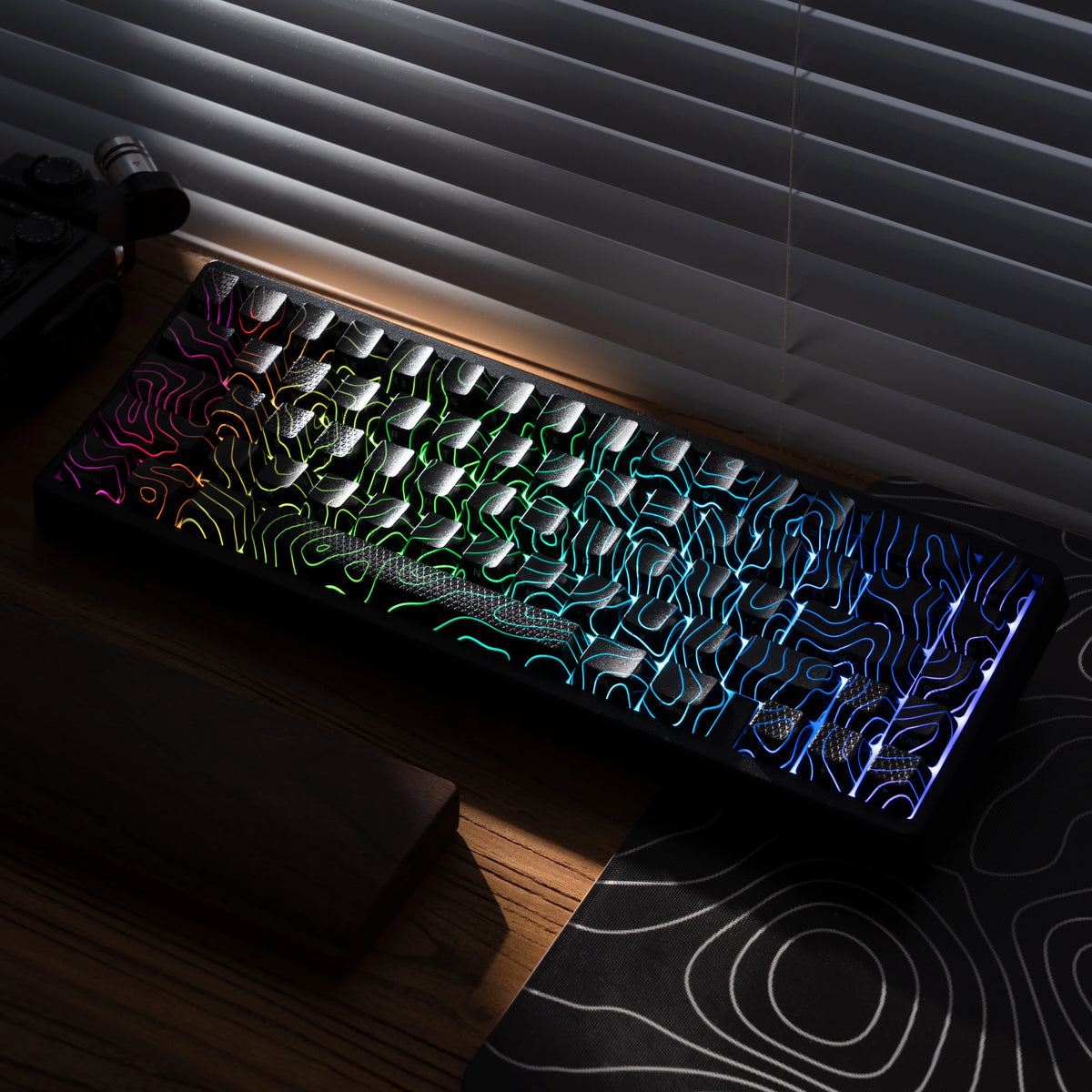 A black mechanical keyboard with colorful RGB lighting is displayed on a wooden surface next to a window blind.