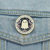 A white ghost enamel pin with floral accents and text on a denim fabric.
