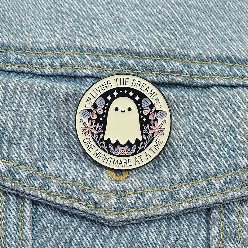 A white ghost enamel pin with floral accents and text on a denim fabric.