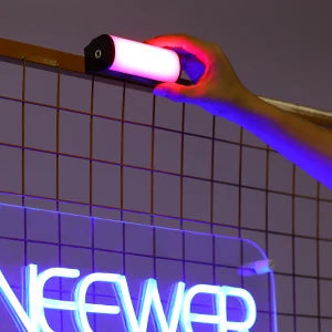 A hand holds a magnetic RGB handheld light wand illuminating a grid background with neon text.